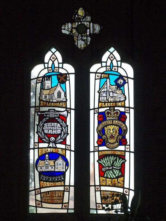 A photo of a stained glass window inside St Leonard's church showing Bretforton institutions, The Church, The Fleece, Bretforton Silver Band, The British Legion, Breforton Village School and a round of asparagus