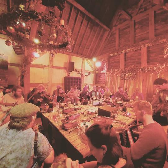 A photo of musicians at a large table playing in the medieval barn of The Fleece Inn Bretforton